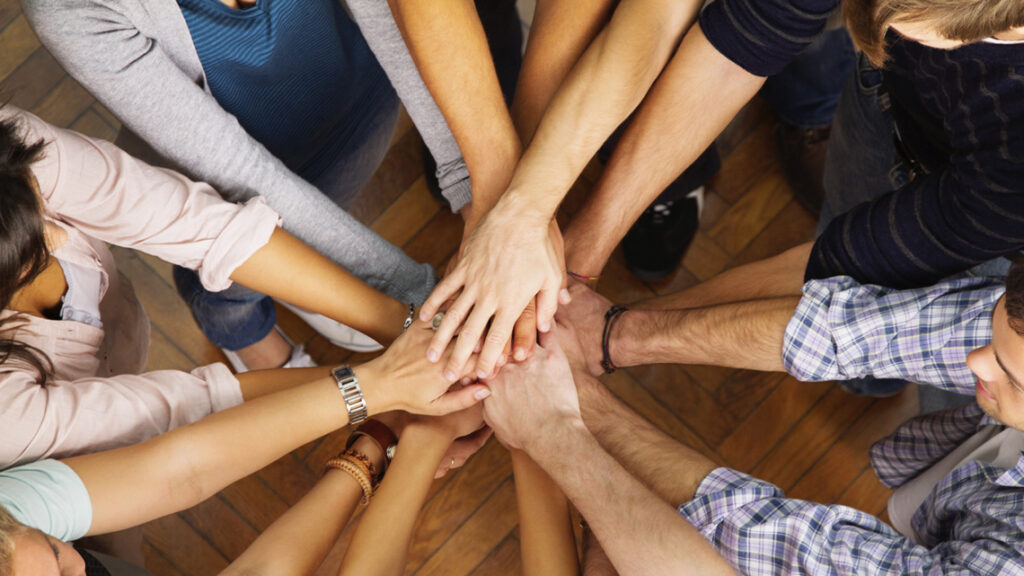 A imagem mostra um grupo reunido em círculo, com as mãos umas sobre as outras, como forma de demonstrar união.